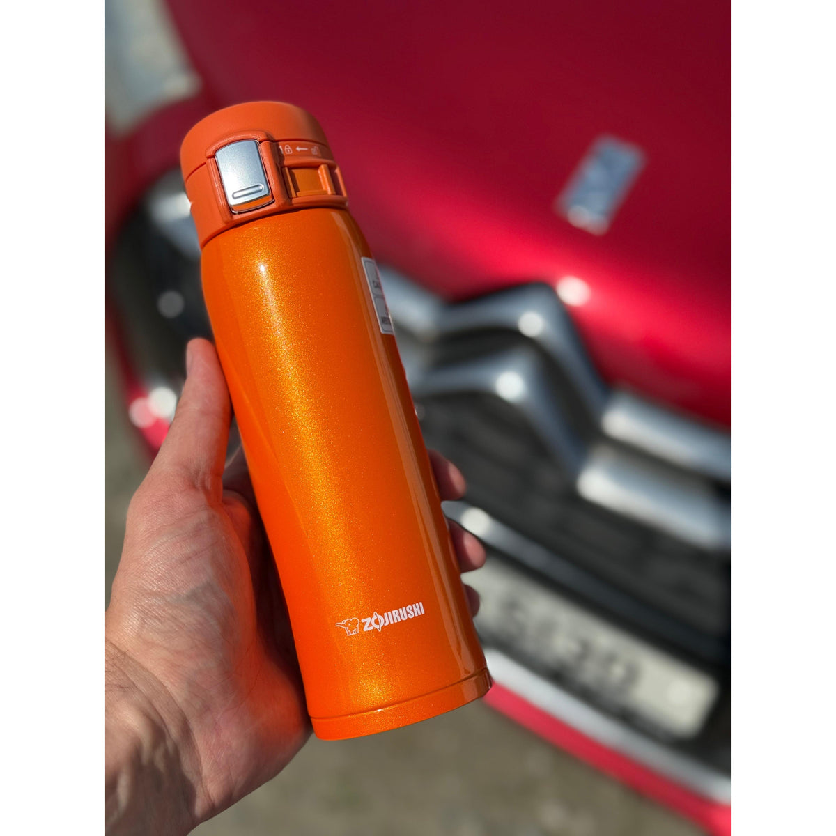 Orange thermos  Zojirushi Mug SM-SHE from 480 ml held by a hand with a red vehicle in the background