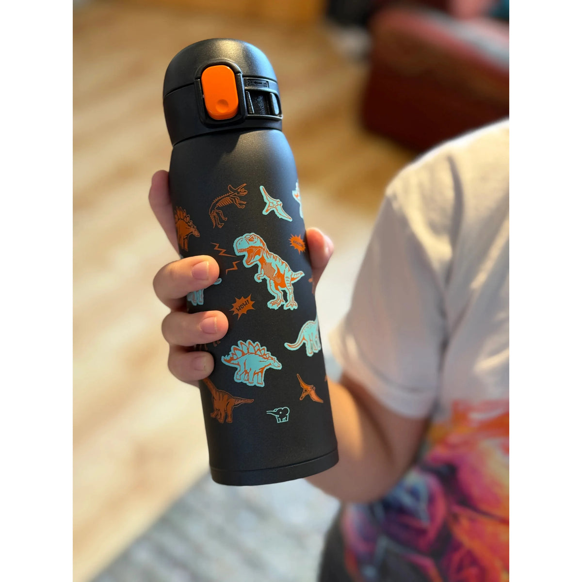 Zojirushi SM-WM48BA kids thermal mug 480 ml (16.20 oz) in black with colourful dinosaur design, reusable insulated travel bottle for hot and cold drinks, held in child’s hand indoors.